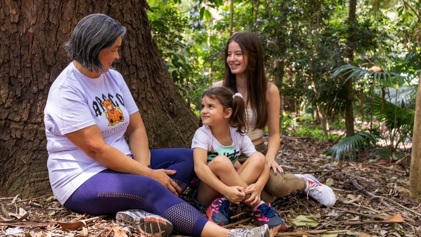 Mulher, adolescente e menina sentadas conversando junto a uma árvore