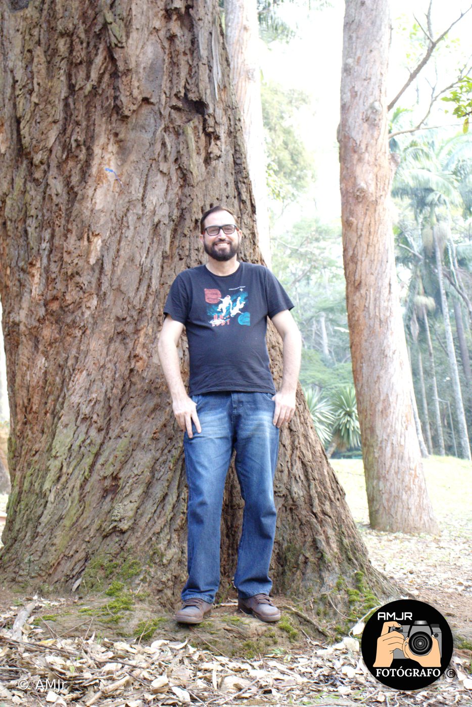 Homem de óculos e camiseta preta em frente a tronco gigante no Horto
